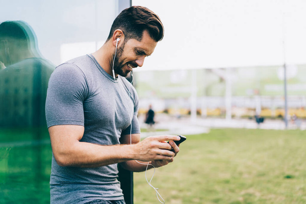 Smiling caucasian guy in active wear and earphones using online app for listening radio via smartphone on morning training, muscular sportsman share media in blog about healthy lifestyle via cellular - Foto, immagini