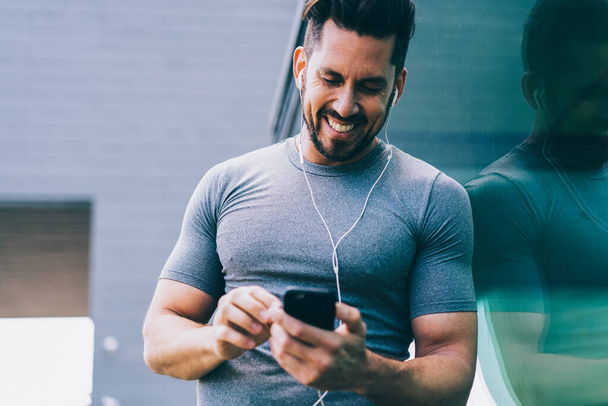 Cheerful caucasian sportsman in earphones satisfied with player on mobile phone choosing music for workout, smiling male in tracksuit using gadgets during pause on training sending mail on cellular - Φωτογραφία, εικόνα