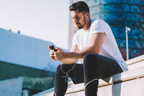 Confident muscular sportsman blogger using smartphone for share media in social networks having break on training, male athlete using earphones for listening music for active lifestyle workout - Zdjęcie, obraz