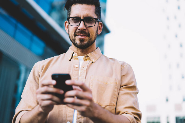 Cheerful 30s male millennial using mobile phone for browsing in social networks standing on street satisfied with 4G connection, prosperous businessman holding cellular typing and sending messages - 写真・画像