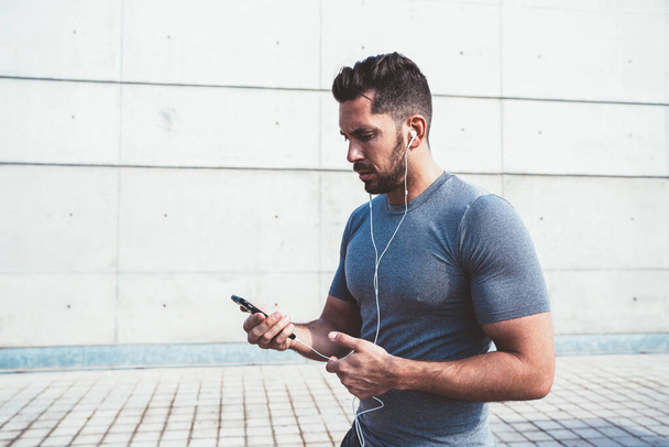 Young 30s caucasian male jogger checking smartphone app notification about cardio workout and calories burning, strong muscular sportsman using mobile phone for listening online radio via earphones - Valokuva, kuva