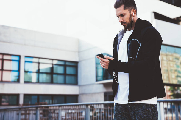 Serious male runner in trendy sports jacket holding mobile phone browse while having workout rest on urban setting, serious caucasian man athlete in earphones listening music and recording results - Fotoğraf, Görsel