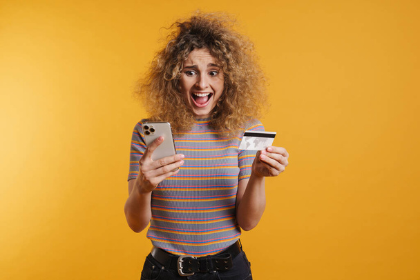 Happy young woman holding mobile phone and credit card celebrating success over yellow wall background - 写真・画像