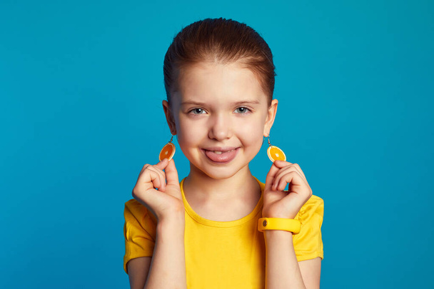 Κορίτσι με κίτρινο πουκάμισο, που δείχνει τη γλώσσα και πορτοκαλί σκουλαρίκια ενάντια στο μπλε τοίχο - Φωτογραφία, εικόνα