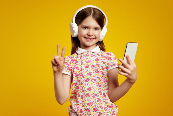 Kind nutzt Smartphone hört Musik über weiße Kopfhörer, zeigt Friedensgeste - Foto, Bild