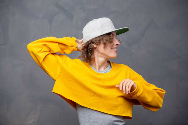 Autumn mood! Portrait of happy smiling woman  with short brunette curly hair wearing yellow sweater and a hat over grey wall background - Fotó, kép
