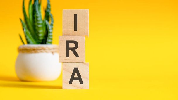 Drei Holzwürfel mit Buchstaben - IRA auf gelbem Tisch, rechts Platz für Text. Konzepte von vorne, Blume im Hintergrund. IRA - kurz für individuelles Rentenkonto - Foto, Bild