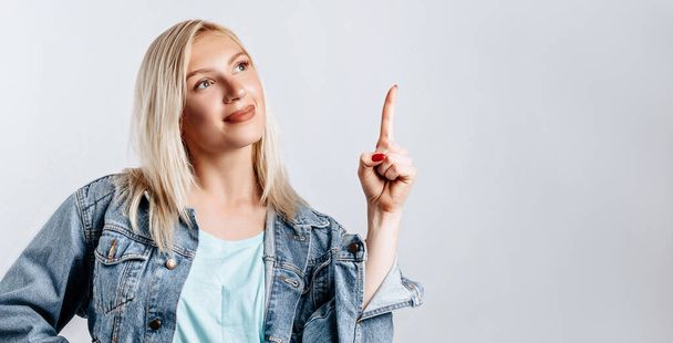 Portrait of a smiling girl pointing finger up at copyspace isolated on a white background. A woman points to an idea, a place for advertising. Positive blonde. - Photo, image