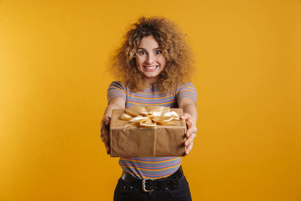 Happy young casual woman holding present box over yellow wall background - Photo, image