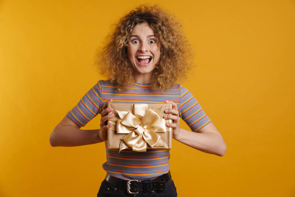 Happy young casual woman holding present box over yellow wall background - Foto, Bild