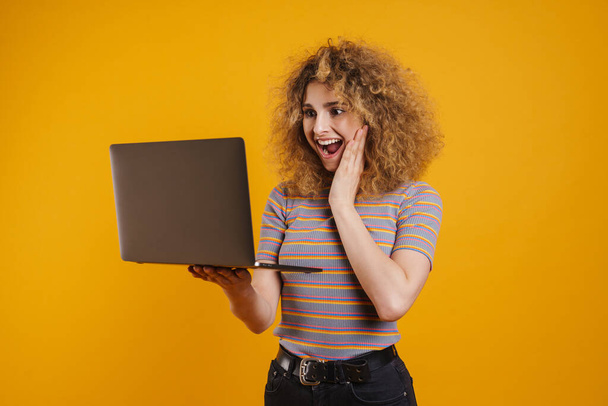 Happy young casual woman holding laptop computer standing over yellow wall background - Фото, изображение
