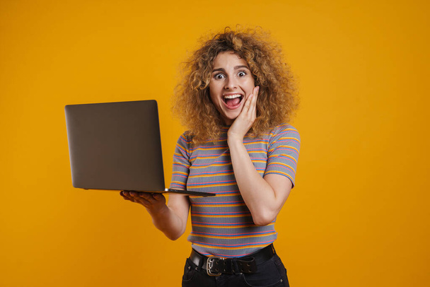 Happy young casual woman holding laptop computer standing over yellow wall background - Photo, image