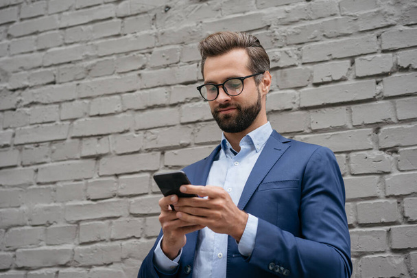 Молодой уверенный бизнесмен держит смартфон, общается онлайн, проверяет электронную почту, стоящую возле серой стены. Красивый бородатый мужчина в стильных очках с помощью мобильного приложения для онлайн-покупок на открытом воздухе  - Фото, изображение