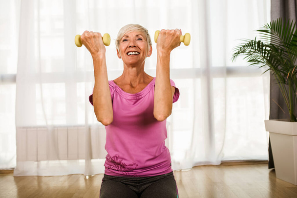 Porträt einer Seniorin, die zu Hause mit Kurzhanteln auf einer Gymnastikmatte sitzt. - Foto, Bild
