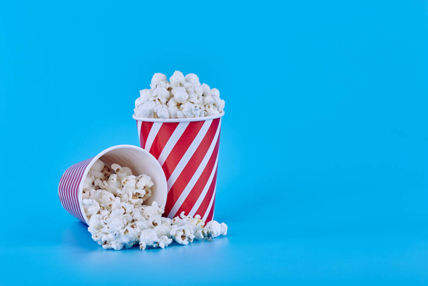 Two glasses full of popcorn on a blue background, one glass upside down and popcorn scattered on a blue background - Foto, Imagen