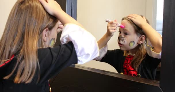 blond zes jaar oud meisje schilderen een roze hart op haar voorhoofd, voor te bereiden op Halloween in gang binnen kijken naar spiegel - Video