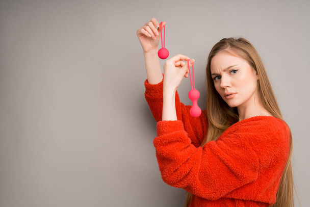 A woman with bright red vaginal Kegel balls in her hands looks with a thoughtful face on a gray background - Fotografie, Obrázek