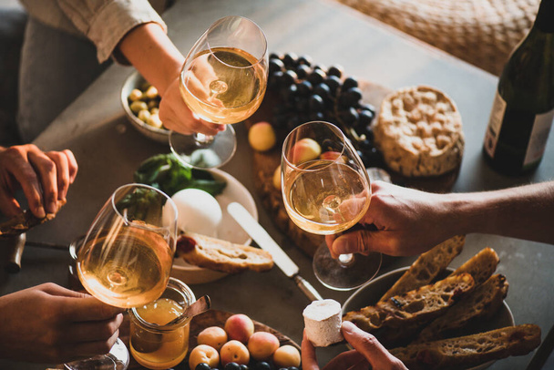 Group of unrecognizable people holding glasses of white orange or rose wine over table with various gourmet snacks cheese grape bread for company. Gathering, celebrating, wine tasting concept - Foto, imagen