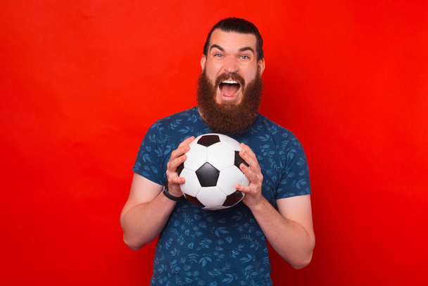 Φωνάζοντας χαρούμενα γενειοφόρος άνδρας κρατά μια μπάλα ποδοσφαίρου με τα δύο χέρια. - Φωτογραφία, εικόνα