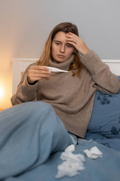 Attractive young woman in beige sweater sitting in bed,  touching head and looking on thermometer. Girl measures body temperature. First symptoms of disease. Female feeling sick, sign of cold and flu. - Photo, Image