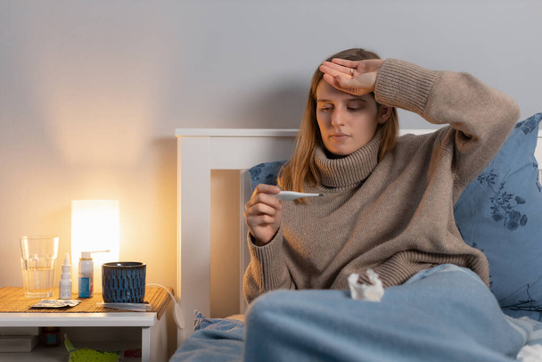 Attractive young woman in beige sweater sitting in bed,  touching head and looking on thermometer. Cute girl measures body temperature. First symptoms of disease. Female feeling cold and flu. - Photo, Image