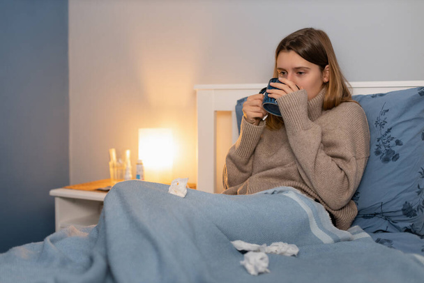 毛布の下のベッドに座っているベージュのセーターのかわいい女の子と熱温度のための救済策を飲む。病気の女。風邪やインフルエンザの症状.季節病だ。鼻を吹くための紙ナプキンの近く. - 写真・画像