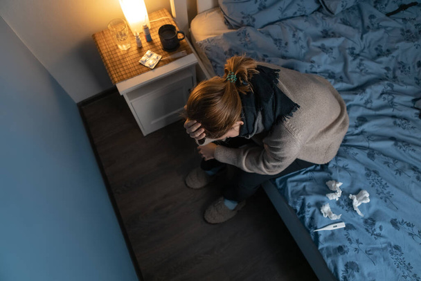 View from above on young girl sitting on the bed and holding her head. Woman feeling sick, suffer from cold and flu. Near paper napkins and thermometer. Female feeling depression. Healthcare concept. - 写真・画像