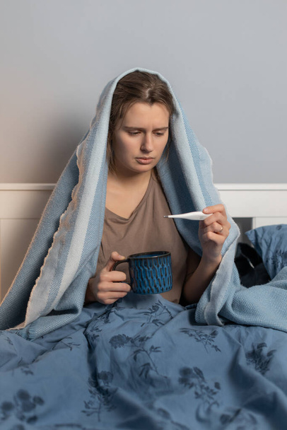 Linda joven envuelta en una manta y mirando un termómetro en la mano. La mujer se siente enferma, calentándose por una taza con bebida caliente. Síntomas de grippe, resfriado o gripe. Concepto sanitario. - Foto, Imagen