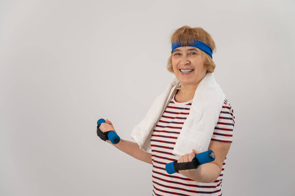 An elderly woman with a blue bandage on her head trains with dumbbells on a white background - Fotó, kép