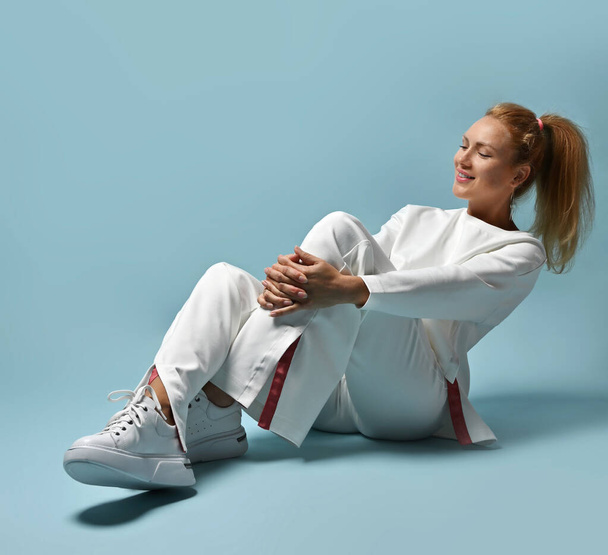 Happy smiling red-haired woman with ponytail hairstyle in white trendy casual pantsuit, kimono style sits on floor - Φωτογραφία, εικόνα