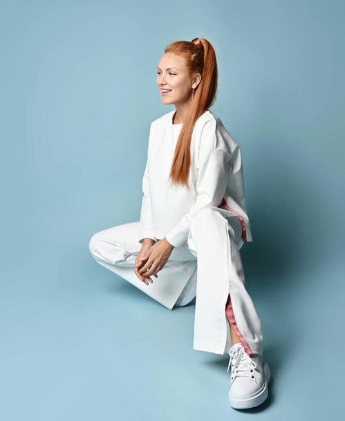Rich redhead young woman in white fashionable pantsuit with pullover, kimono style sits on her haunches - Zdjęcie, obraz