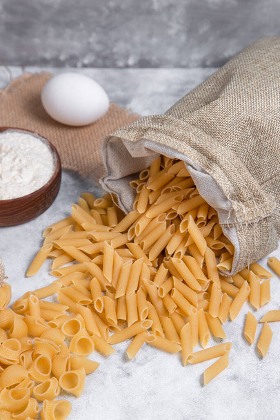Sack bags of dried penne pasta and shell shaped macaroni with a wooden bowl of flour . High quality photo - Photo, Image