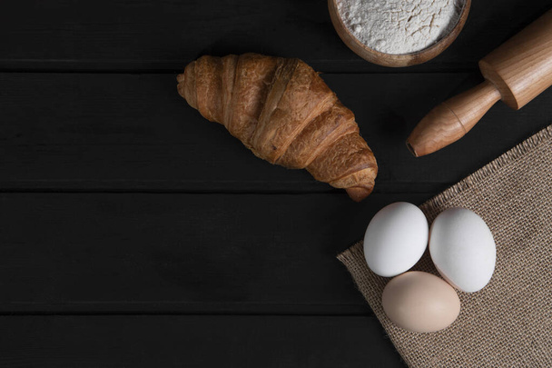 Plain croissants, bowl of flour, rolling pin and raw eggs on dark wooden surface. High quality photo - Photo, Image