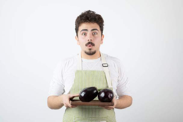 Jeune cuisinier masculin dans un tablier tenant une assiette de grosses aubergines sur fond blanc. Photo de haute qualité - Photo, image