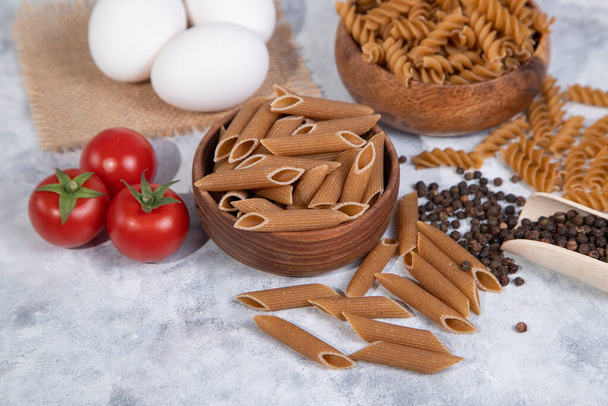 Un bol en bois de pâtes italiennes sèches non cuites Penne rigate avec des tomates et des grains de poivre. Photo de haute qualité - Photo, image