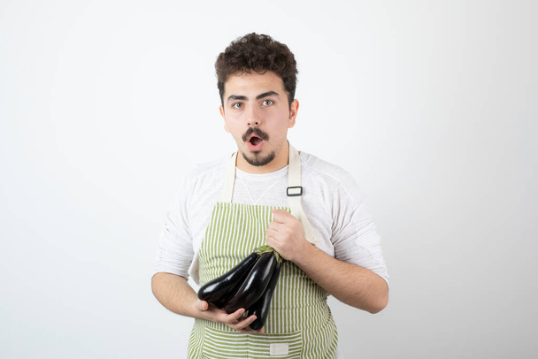 Photo d'un jeune homme surpris dans un tablier tenant deux grosses aubergines fraîches. Photo de haute qualité - Photo, image