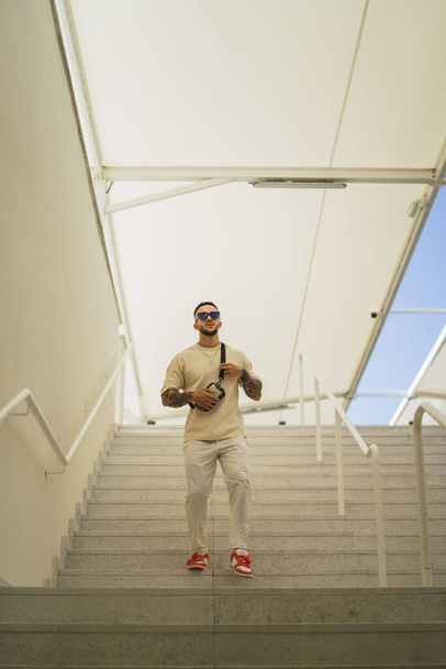 A vertical shot of a young caucasian stylish guy with tattoos and sunglasses standing on the stair - Фото, изображение