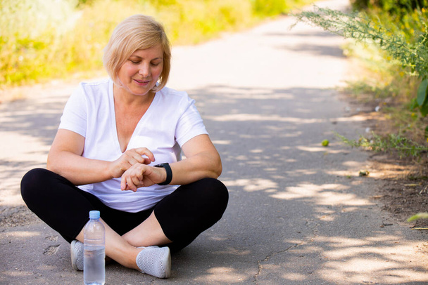 The photo of a happy blonde sporty woman sitting in a lotus position on the road in the park wearing sports clothes pointing on the digital watch on her hand. - Fotoğraf, Görsel