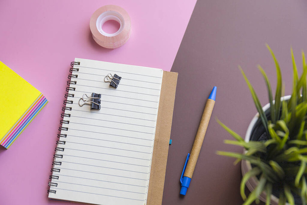 A top view shot of a notebook, sticky paper,tape, pen, and a plant on a pink and brown background - Photo, image