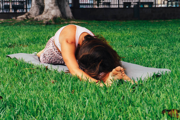 A young Spanish woman doing the hanumanasana yoga pose on a mat in a park - 写真・画像