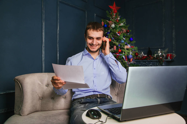 Un tipo con una camisa azul se sienta en un portátil y habla por teléfono contra el fondo de un árbol de Navidad. Un hombre trabajando en una computadora durante las vacaciones de Navidad - Foto, Imagen