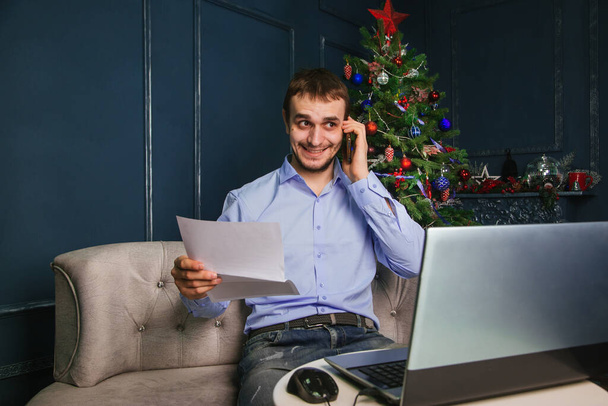 A guy in a blue shirt sits at a laptop and talks on the phone against the background of a Christmas tree. A man working at a computer during the Christmas holidays - Photo, Image