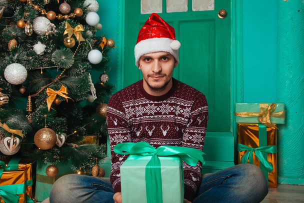Der Typ im weinroten Pullover im skandinavischen Stil im Neujahrsinterieur. Ein Mann mit Weihnachtsmütze hält eine Geschenkbox in der Hand. Eine Überraschung wartet - Foto, Bild