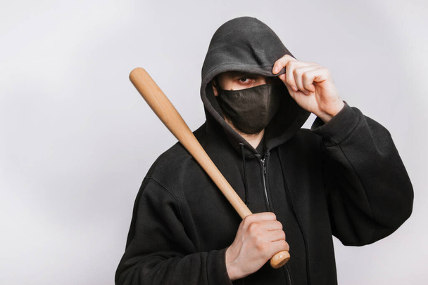 Een gemaskerde bandiet in een zwarte hoodie, jeans en een knuppel op een witte achtergrond. De crimineel bedreigt - Foto, afbeelding