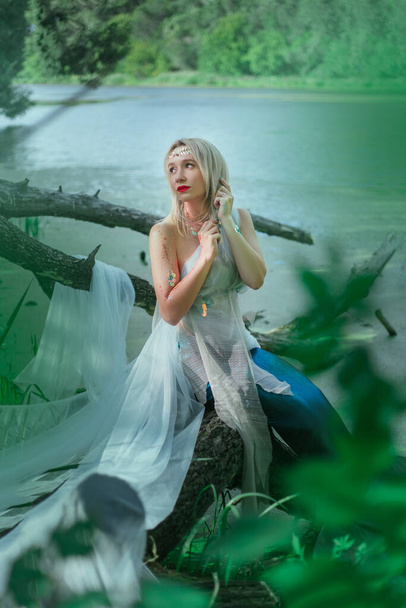 Una hermosa chica en un paño blanco transparente se sienta en una rama en un pantano. Una sirena con una diadema se sienta en un árbol junto a un depósito verde. - Foto, imagen