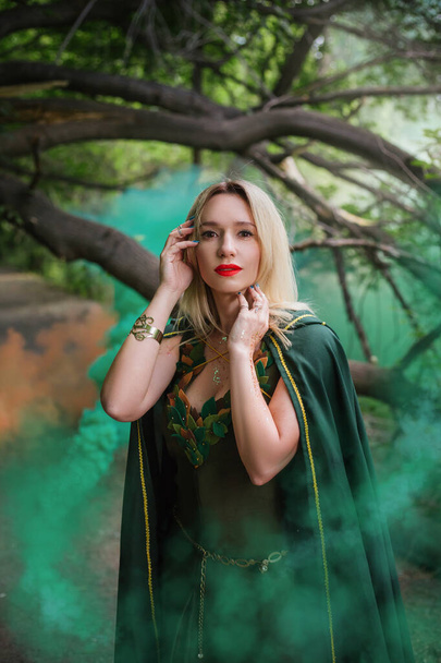 Een blonde in een groene mantel van een druïde en in een fantastische jurk met bladeren in de mist. Een tovenares in middeleeuwse jurk staat in het bos in groene en oranje rook. Het beeld van een goede heks - Foto, afbeelding