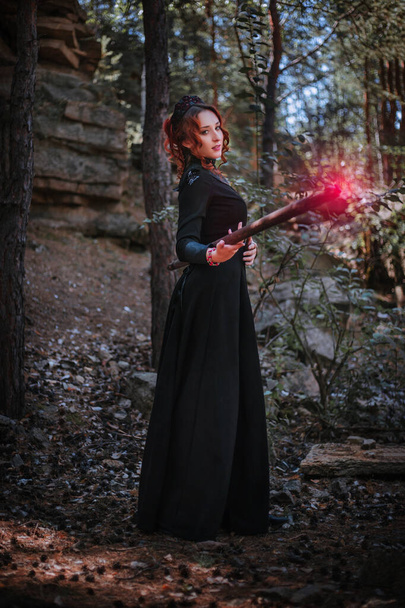 Una bruja con un vestido largo negro del estilo eslavo sostiene un bastón en sus manos. Una mujer evoca en un bosque oscuro. - Foto, Imagen