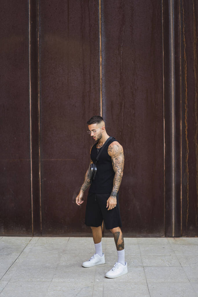 A stylish caucasian guy with cool tattoos standing on the paved road on the brown background - Fotografie, Obrázek