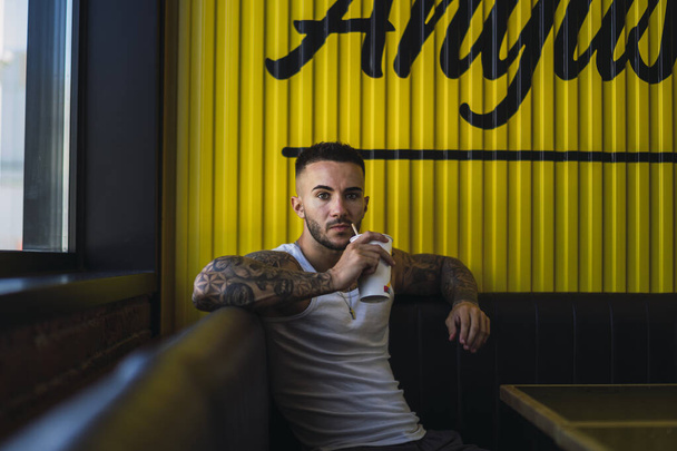 A young caucasian guy with cool tattoos sitting in the cafe and drinking a cup of juice - Fotoğraf, Görsel
