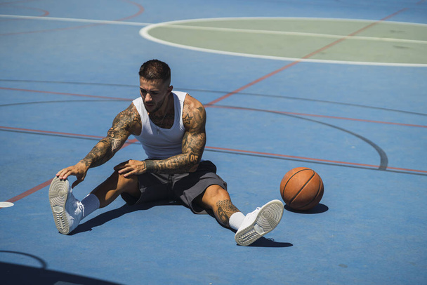 A young caucasian guy with cool tattoos sitting on the basketball court with a ball doing exercises - Foto, Imagen